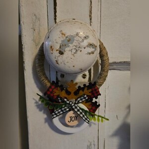 以下が含まれることがあります： 白いドアノブに茶色のロープのリースが取り付けられています。リースは茶色の雪片、赤と黒の格子縞の布、白黒のリボン、緑の葉で飾られています。リースから「JOY」と書かれた白い円がぶら下がっています。