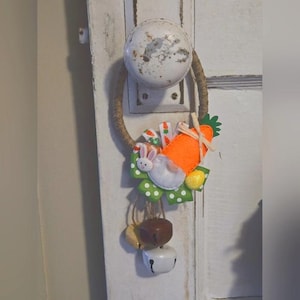 Könnte beinhalten: Ein handgemachter Oster-Türkranz mit einer Karotte, einem Hasen und einer grünen Schleife. Der Kranz ist aus Schnur gefertigt und hat drei Glöckchen, die daran hängen.