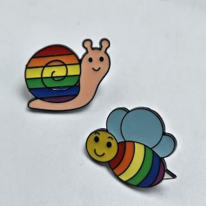 Op de afbeelding: Twee emaille pins: een slak met een regenboogschelp en een lachende bij met regenboogstrepen en blauwe vleugels. Beide pins hebben zwarte contouren en staan tegen een witte achtergrond. De slak is lichtroze en de bij heeft een gele kop.
