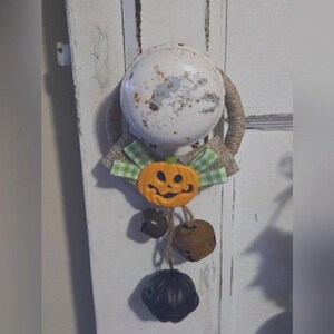 Könnte beinhalten: Ein weißer Türknauf mit einer Schleife aus Sackleinen und einem grün-weiß karierten Band. An der Schleife ist ein kleiner orangefarbener Kürbis mit einem Jack-o'-Lantern-Gesicht befestigt. Drei braune Glöckchen und eine schwarze Glocke hängen an dem Kürbis.