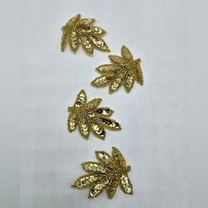 May include: Four gold leaf-shaped appliques. Each applique features a detailed design with gold sequins and beads, creating a shimmering effect. The appliques are arranged on a white surface, showcasing their intricate craftsmanship.