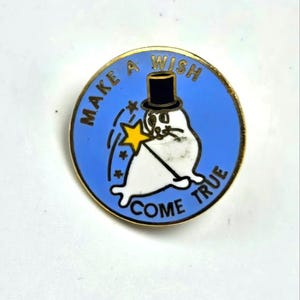 May include: A round pin with a blue background and gold trim. The pin features a white seal wearing a black top hat and holding a yellow star wand. The text "MAKE A WISH COME TRUE" is in gold.