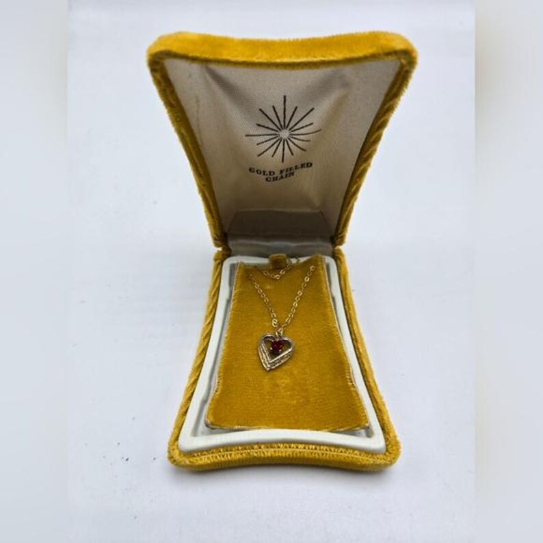 May include: A gold-filled chain necklace with a heart-shaped pendant featuring a red stone in the center. The necklace is displayed in a yellow velvet box with the words "Gold Filled Chain" printed on the inside.