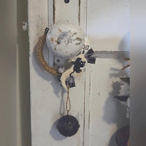 Pode incluir: Uma porta branca com uma guirlanda de Halloween de juta com um gato preto e uma teia de aranha pendurada na maçaneta. A guirlanda é feita de juta e tem um sino preto pendurado.
