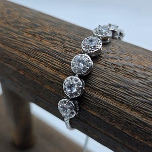 May include: A silver bracelet with round, clear gemstones. The bracelet features a series of bezel-set stones, each surrounded by smaller accent stones. The bracelet is displayed on a wooden surface.