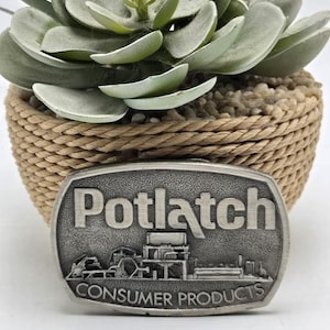 May include: A silver-toned belt buckle featuring the word "Potlatch" in relief. Below, the words "Consumer Products" and an industrial design are visible. The buckle is positioned in front of a decorative plant in a woven basket.