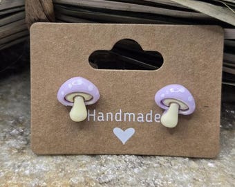 May include: A pair of purple mushroom stud earrings with white polka dots and a yellow stem. The earrings are displayed on a brown card with the word "Handmade" written on it.