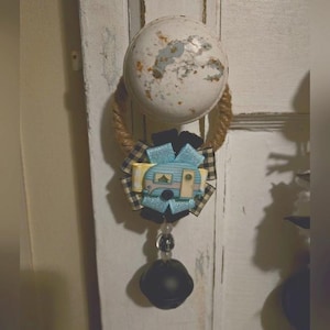 Puede incluir: Un amuleto de caravana azul y blanco con una cinta a cuadros blanco y negro y una campana negra colgando de un lazo de cuerda.