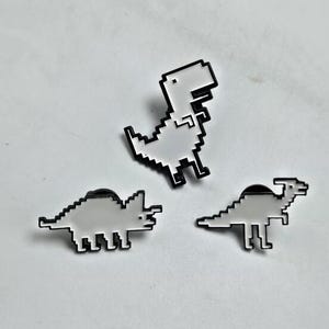 May include: Three pixelated dinosaur pins in black and white. The pins feature a Tyrannosaurus Rex, a Triceratops, and a Parasaurolophus. The pins are set against a white background.