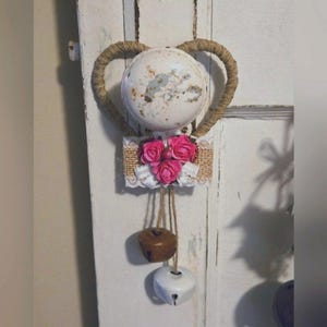 May include: A decorative heart-shaped wall hanging. A distressed white metal sphere is framed by a jute rope heart. Below, a burlap and lace accent features pink roses. Two bells hang beneath, one brown and one white.
