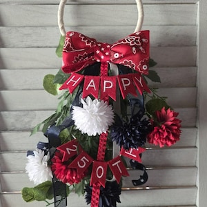 May include: A decorative wall hanging featuring a white circular hoop, a red bandana-print bow, and red felt pennants spelling HAPPY 25TH. The arrangement includes white, red, and dark blue artificial flowers, green foliage, and dark blue ribbons.