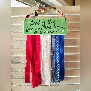 Op de afbeelding: Een patriottische wanddecoratie met een groen bord met de tekst "land of the free and the home of the brave". Rode, witte en blauwe linten hangen onder het bord, met decoratieve elementen aan de bovenkant.