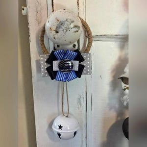 Puede incluir: Un pomo de puerta decorativo con un adorno de lazo y campana. El lazo presenta cintas negras, azules y blancas con un diseño de insignia. Una campana blanca con una estrella y un diseño recortado cuelga debajo.