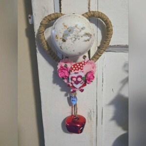 Op de afbeelding: Een decoratief hartvormig ornament met een witte, verweerde deurknop aan de bovenkant. Het hart is omwikkeld met touw en versierd met roze rozen en een kabouterfiguur. Een blauw hart met het woord "LOVE" hangt eronder, met een rode bel aan de onderkant.