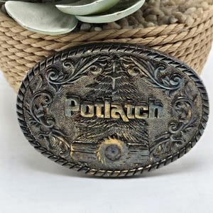 May include: An oval, antique-style belt buckle with the word "Potlatch" in raised lettering. The buckle has an ornate, engraved design with a central image of a tree and a saw. The color is a mix of silver and bronze.