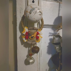 Puede incluir: Un colgador de puerta rústico con un diseño de arpillera y girasol. Presenta una pequeña aplicación de pollo y tres campanas. El colgador está sujeto a una puerta blanca con un pomo de puerta vintage.