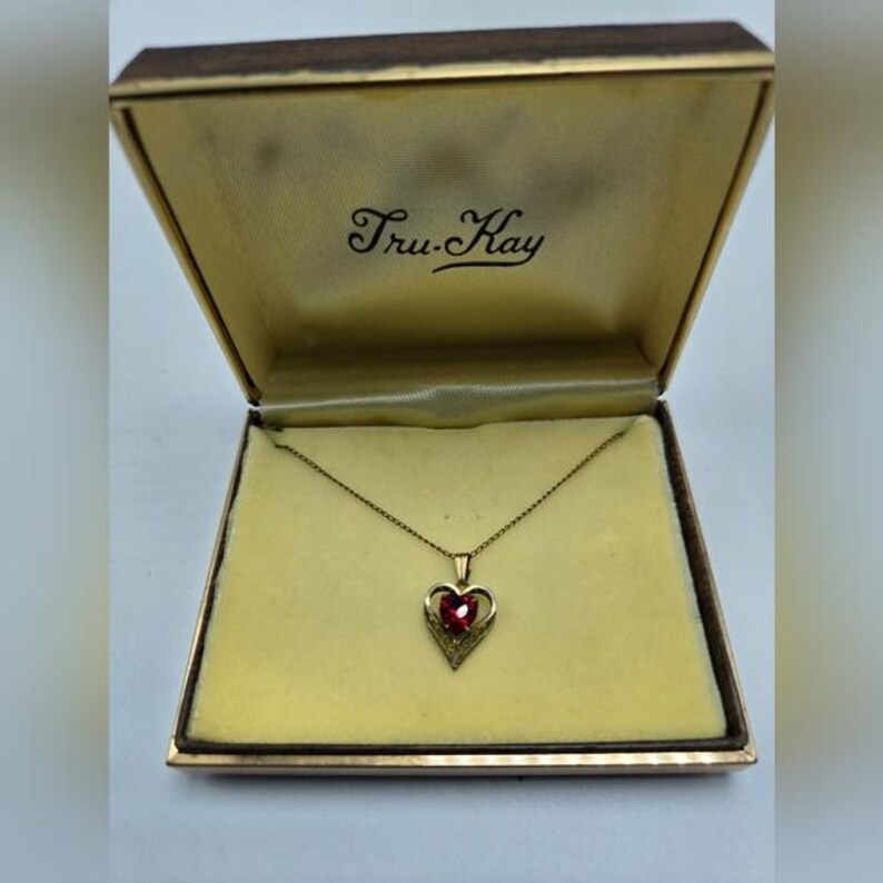 May include: A gold heart-shaped pendant necklace with a red gemstone in the center, in a Tru-Kay jewelry box.