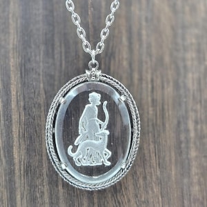 May include: Silver-toned pendant necklace featuring an oval, clear glass pendant with a carved design of a figure with a bow and a dog. The pendant is framed with a twisted rope design and hangs from a delicate chain. The background is a wooden surface.