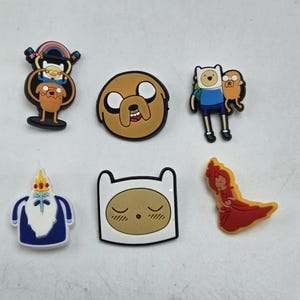 May include: A selection of Adventure Time cartoon character charms. The charms depict characters including Finn, Jake, and Princess Bubblegum. These colourful charms are made from a rubbery material and are designed to be attached to shoes or other accessories.