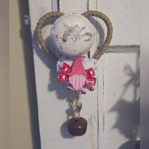 Puede incluir: Una decoración de gnomo para el Día de San Valentín con un marco en forma de corazón hecho de cuerda, un gorro de gnomo rosa y blanco y un cuerpo a rayas rosa y blanco. El gnomo sostiene una pequeña campana con un amuleto en forma de corazón.
