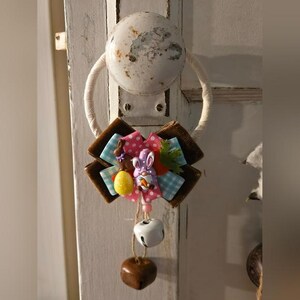 Puede incluir: Adorno de puerta con temática de Pascua. El lazo decorativo presenta un diseño de conejito y huevo, con lunares rosas, cuadros azules y una pequeña campana y un dije en forma de corazón. La puerta es blanca.