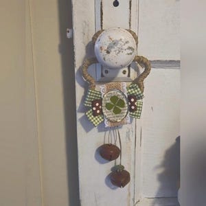 Puede incluir: Adorno para pomo de puerta con un pomo blanco envejecido y un asa de cuerda de yute. Incluye un lazo a cuadros verde y blanco, un trébol de cuatro hojas y dos pequeñas campanas marrones. El diseño tiene una estética rústica y campestre.