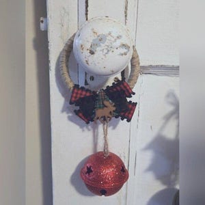May include: A red glitter bell with a plaid bow and a moose charm hangs from a rope loop attached to a white door knob.