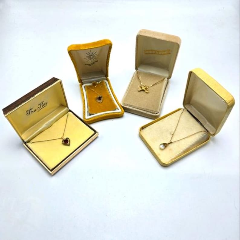 May include: Four small, yellow velvet jewelry boxes with gold necklaces inside. The boxes are on a white surface. The necklaces have different pendants, including a heart, a teardrop, and a butterfly.