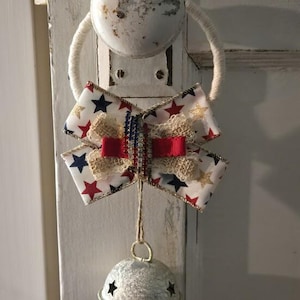 May include: A decorative door knocker featuring a patriotic bow with red, white, and blue star patterns, burlap, and a red ribbon. A silver bell with star cutouts hangs below. The knocker is attached to a white, distressed door.