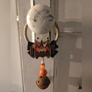 Könnte beinhalten: Ein Türklopfer zum Thema Halloween mit einem weißen, runden Griff und einer Schlaufe aus Juteseil. Eine schwarze Schleife mit einem "Happy Halloween"-Schild und eine Dekoration aus Zuckermais und Glocke hängt darunter.