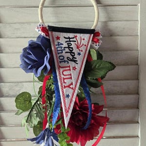 Op de afbeelding: Een decoratieve wandhanger met een witte viltwimpel met de tekst Happy 4th of July in zwart en rood, geaccentueerd door blauwe sterren en rood vuurwerk. Het hangt aan een crèmekleurige ring en is versierd met rode en blauwe stoffen bloemen.