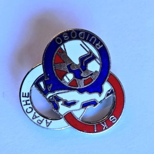 Puede incluir: Pin con tres círculos entrelazados en blanco, rojo y azul. El círculo superior dice "RUIDOSO" en azul, el inferior "SKI" en rojo y el izquierdo "APACHE" en blanco. El centro representa un esquiador.