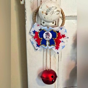 May include: A patriotic door hanger featuring a white door knob with a rope loop. A bow with red, white, and blue stars and a central emblem that says "Trump Girl" is attached. A red bell with star cutouts hangs below.