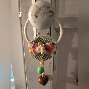 May include: A decorative Easter-themed hanging ornament. It features a white rope loop attached to a white and distressed knob. The ornament includes a bow with a bunny, flower, and green bell, and a brown acorn-shaped element.