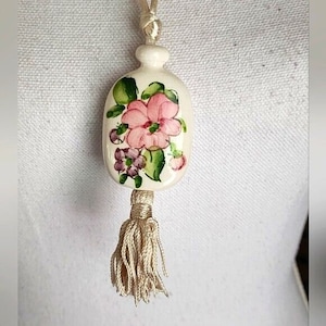 May include: A white ceramic pendant with a floral design. The pendant features a pink flower with green leaves and a white tassel.