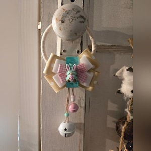 Puede incluir: Un lazo decorativo con temática de Pascua con un motivo de conejito. El lazo presenta cintas blancas, rosas y amarillas con lunares. Una pequeña figura de conejito está centrada en una cinta turquesa. Una cadena de cuentas y una campana cuelgan debajo.