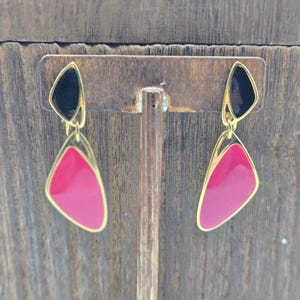 May include: A pair of vintage earrings with a gold-tone frame. Each earring features a black upper section and a pink, tear-drop shaped lower section. The earrings are displayed on a wooden stand against a wooden background.