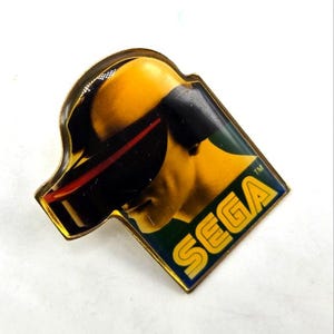 Puede incluir: Un pin SEGA vintage con una imagen estilizada de una persona con gafas futuristas. El pin tiene un borde dorado y el logotipo de SEGA en amarillo. El fondo es una mezcla de amarillo, negro y verde.