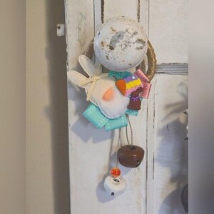 May include: A white felt bunny with a pink nose and blue ribbon hangs from a rusty metal ring with a brown and gold rope. The bunny is holding a carrot and a small chocolate bunny.