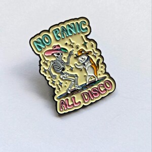 Op de afbeelding: Emaille pin met een skelet en een kat met hoeden, dansend op een surfplank. De pin heeft de tekst "NO PANIC ALL DISCO" in een bubbellettertype met een gele achtergrond en zwarte rand.