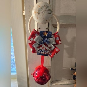 May include: A patriotic door hanger featuring a red, white, and blue bow with a star and stripes pattern. A red bell hangs below, and the number "250" is visible on the bow. The hanger is attached to a white door knob.