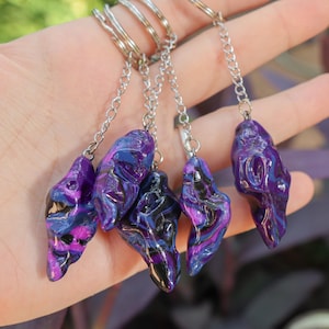 May include: Four keychains featuring irregular, organic-shaped pendants with a glossy, marbled finish in shades of deep purple, black, and bright magenta. Each pendant is attached to a silver-toned metal chain and a circular split ring for keys.