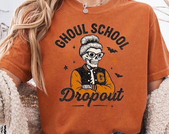 May include: Orange t-shirt with the text "Ghoul School Dropout" and a skeleton graphic wearing glasses and a letterman jacket. The shirt is a fall-themed design, perfect for Halloween or spooky season.