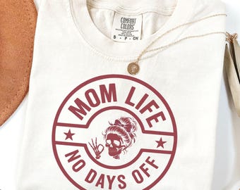 May include: Ivory-colored t-shirt with a round, burgundy graphic that reads "MOM LIFE NO DAYS OFF." The graphic features a skull with a bun and a hand making the okay sign. A gold necklace and a brown hat are also visible.
