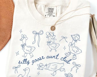 May include: Ivory-colored t-shirt with a blue line drawing of geese wearing hats and bows. The text "silly goose aunt club" is printed in blue. A gold necklace and a brown hat are also visible.