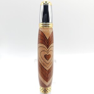 May include: A wooden pen with a heart design. The pen is made of different types of wood, including maple, walnut, and cherry. The pen has a gold-colored metal top and bottom.