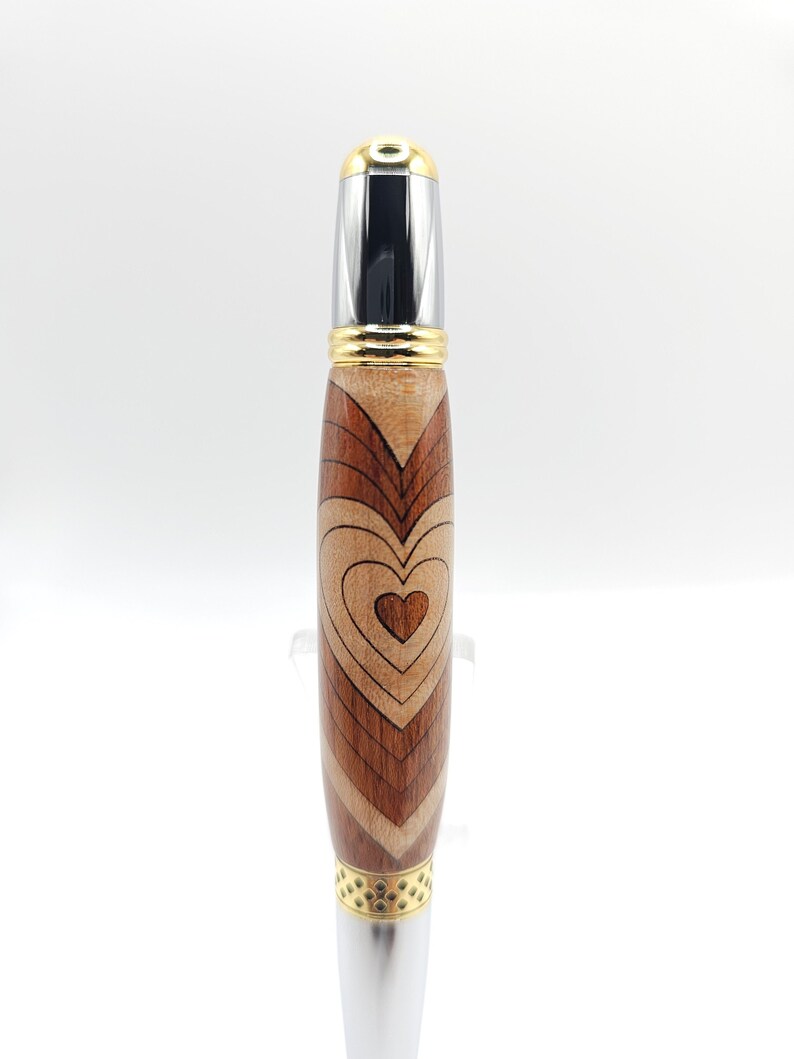 May include: A wooden pen with a heart design. The pen is made of different types of wood, including maple, walnut, and cherry. The pen has a gold-colored metal top and bottom.