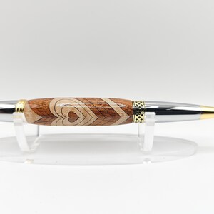 May include: A wooden pen with a silver and gold finish. The pen has a heart design and is on a clear acrylic stand.