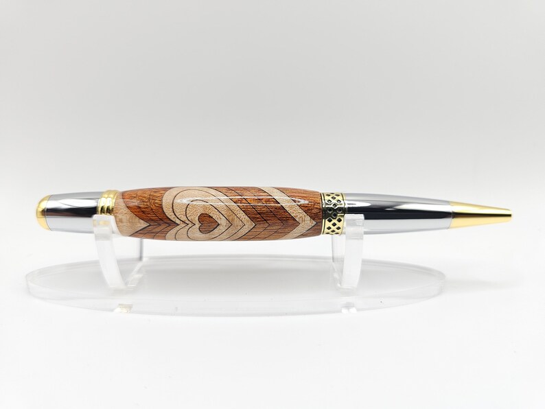 May include: A wooden pen with a silver and gold finish. The pen has a heart design and is on a clear acrylic stand.