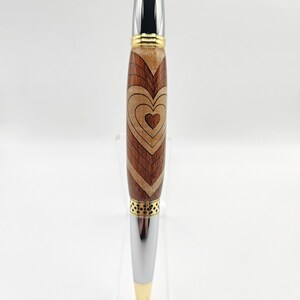 May include: A wooden pen with a heart design in shades of brown and gold. The pen has a silver metal tip and a gold band.
